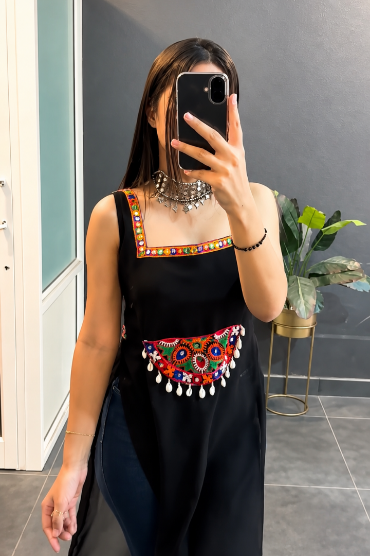A woman takes a mirror selfie wearing a black sleeveless dress with colorful embroidered details on the neckline and waist, featuring dangling white tassel accents. She is standing in an indoor setting with a gray wall, a glass door, and a potted plant in the background.