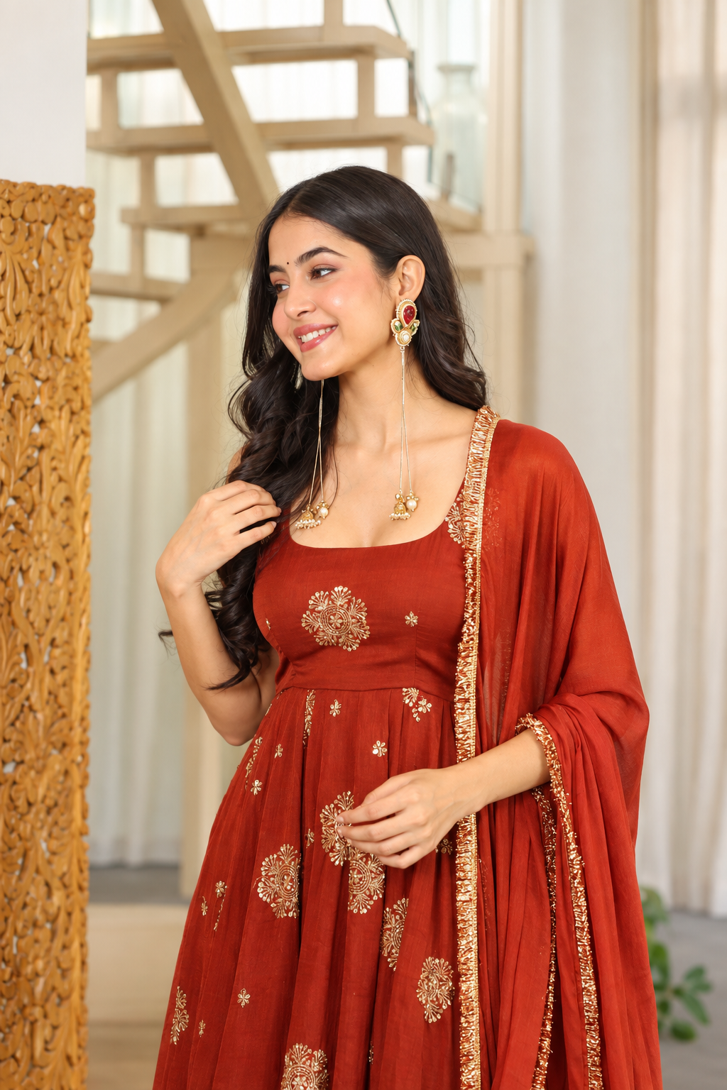 A woman wearing a rust-orange Anarkali gown with gold floral embroidery and a matching dupatta draped over her shoulder, standing in an indoor setting with a staircase in the background.