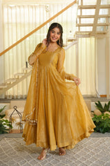 A woman models a long, flowing mustard yellow traditional dress with sheer sleeves and decorative beadwork, standing in a bright, modern indoor setting with a staircase and plants in the background.