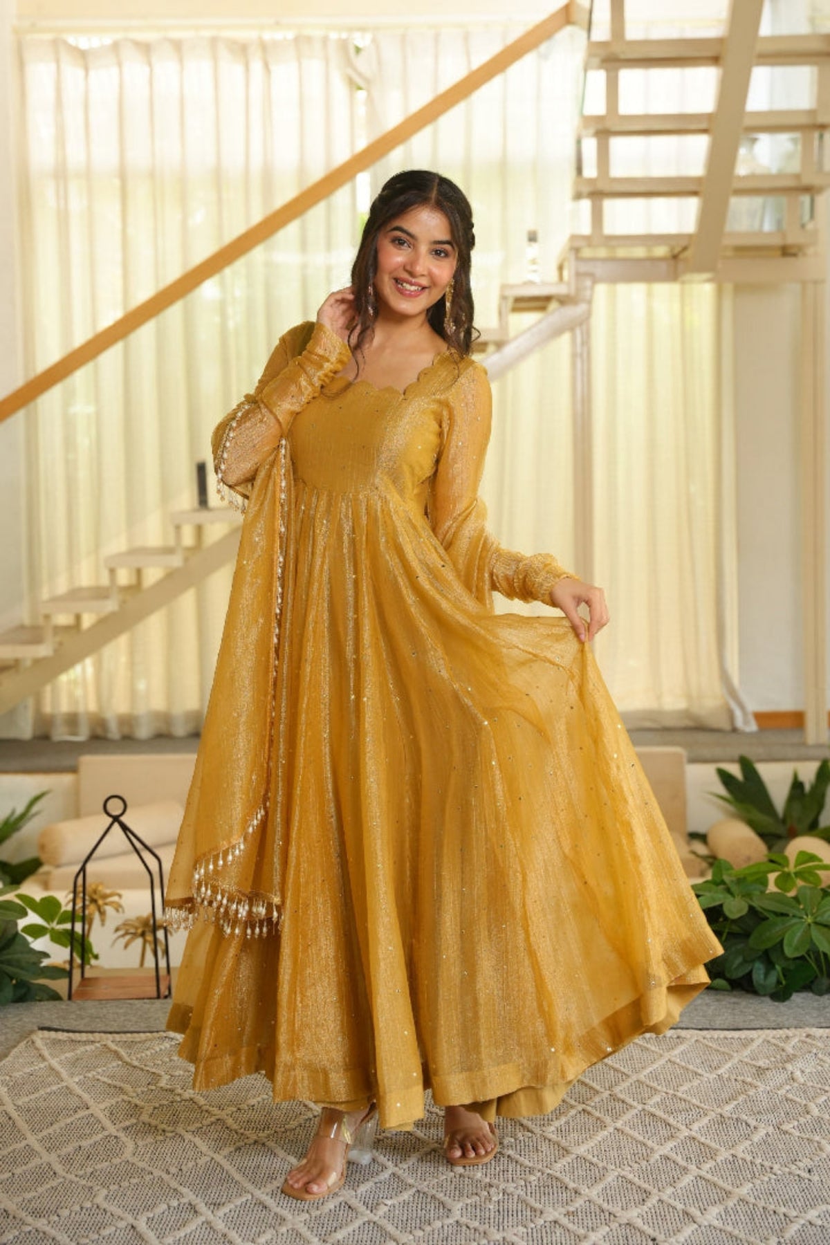 A woman models a long, flowing mustard yellow traditional dress with sheer sleeves and decorative beadwork, standing in a bright, modern indoor setting with a staircase and plants in the background.