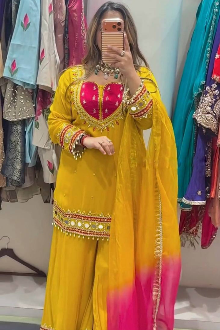 A woman takes a mirror selfie wearing a bright yellow traditional outfit with a pink and yellow gradient dupatta, decorated with gold mirror work and embroidery. The background shows a clothing store with other colorful garments hanging on racks.