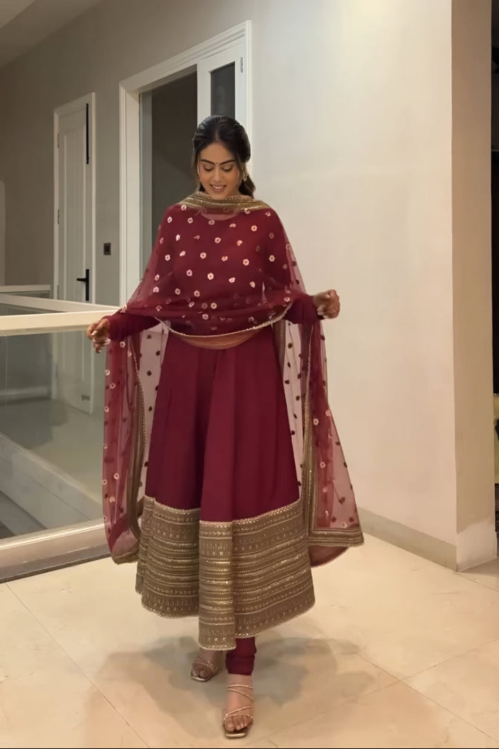 Woman in a maroon traditional outfit with gold details in a home setting.