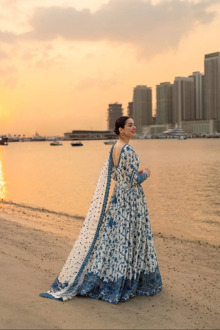 Woman in a blue and white traditional outfit standing on a beach with city skyline in the background.