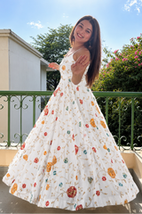 A woman wearing a white sleeveless maxi dress with a colorful floral print stands on a balcony, holding out her hand as if to wave. The dress features a fitted bodice and a voluminous, flowing skirt.