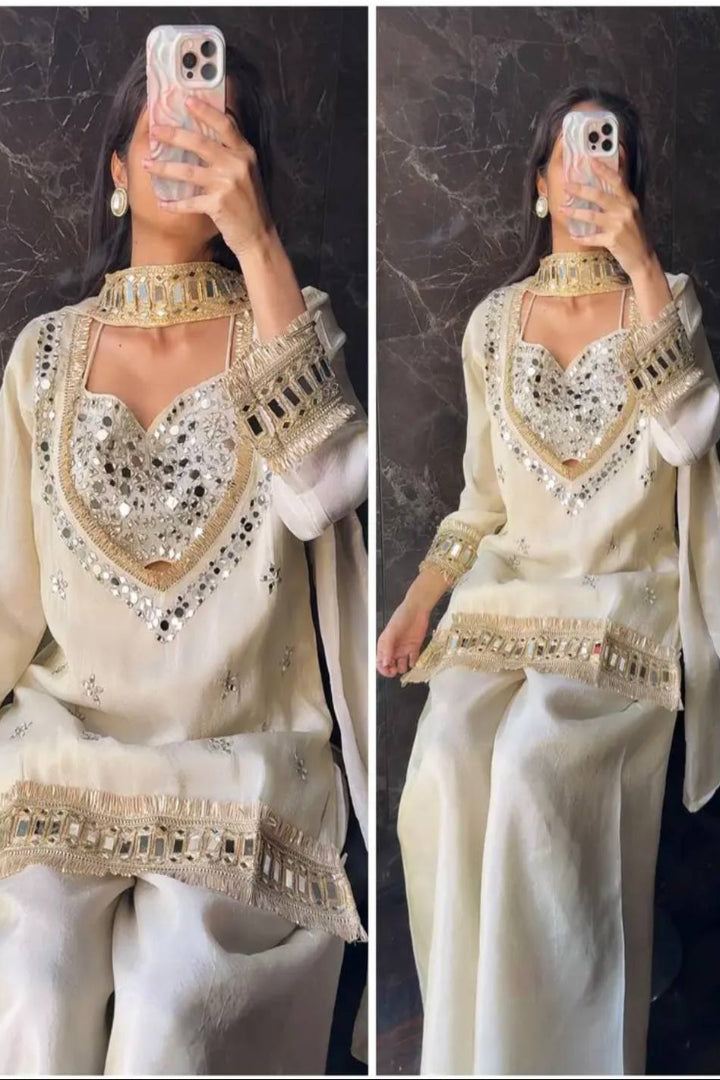 Woman wearing a white traditional outfit with gold embroidery, sitting against a dark background.
