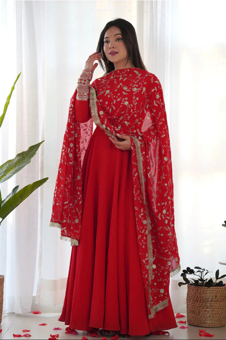 Woman in a red traditional outfit with gold patterns standing in a bright room with white curtains and plants.