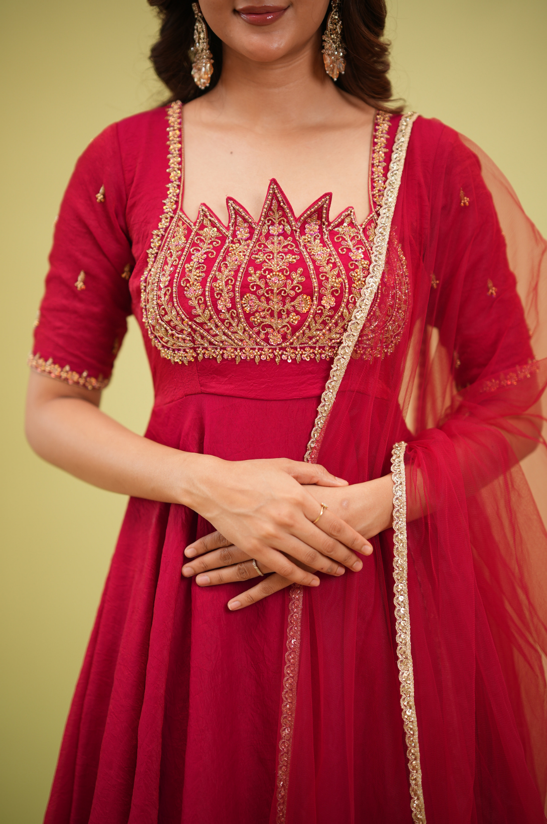 A woman wearing a bright red Indian ethnic gown with elaborate gold embroidery on the bodice and sleeves, paired with a matching sheer dupatta, set against a solid pale yellow background.
