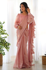 Woman wearing a pink saree in a bright room with white curtains and a plant.