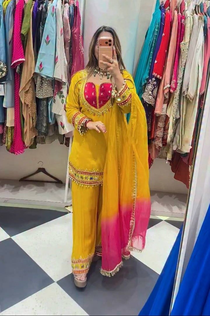 A woman takes a mirror selfie in a clothing boutique wearing a bright yellow traditional outfit with a pink gradient dupatta and a pink embroidered bodice. Clothing racks filled with colorful garments are visible in the background.