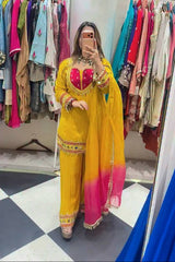 A woman takes a mirror selfie in a clothing boutique wearing a bright yellow traditional outfit with a pink gradient dupatta and a pink embroidered bodice. Clothing racks filled with colorful garments are visible in the background.
