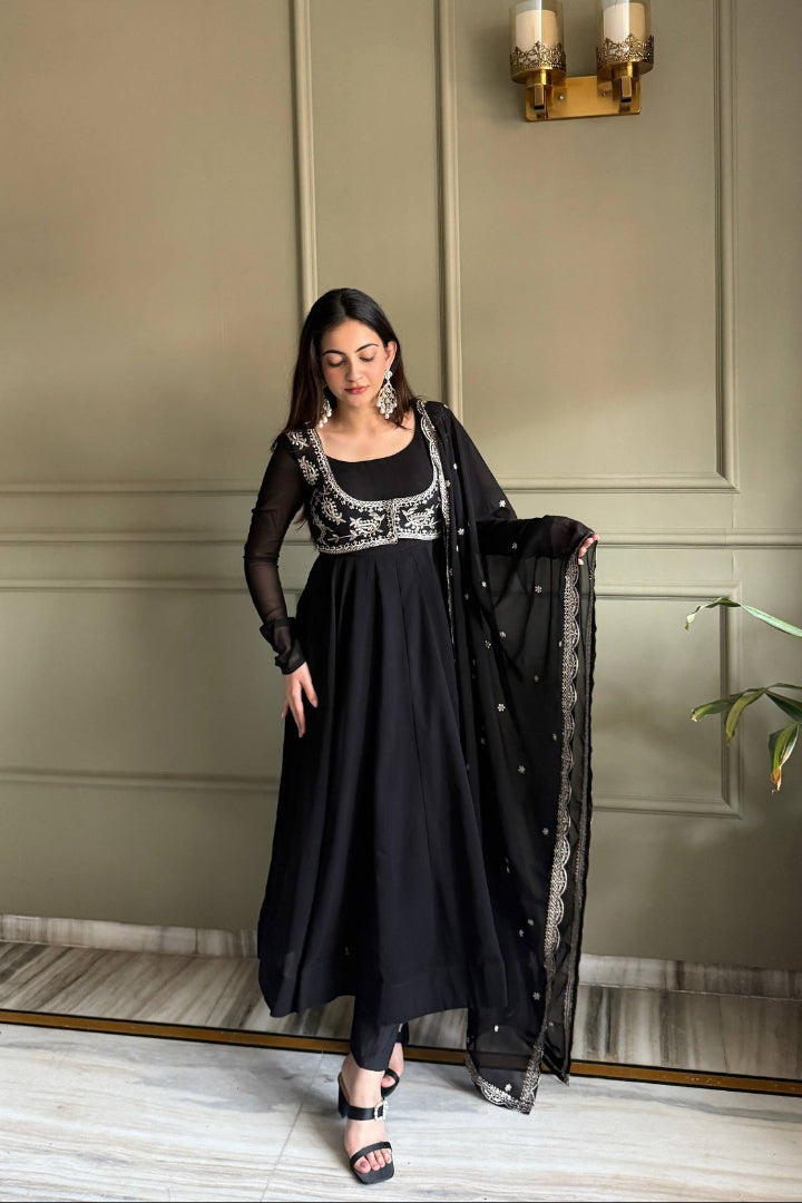 Woman in a black traditional outfit with silver embroidery standing against a neutral wall.