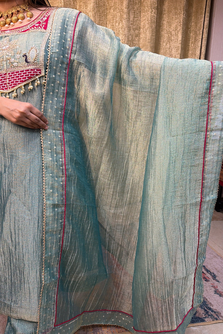 A close-up of a light blue-green sheer fabric garment with pink border trim and gold embroidered detailing, shown on a person indoors.