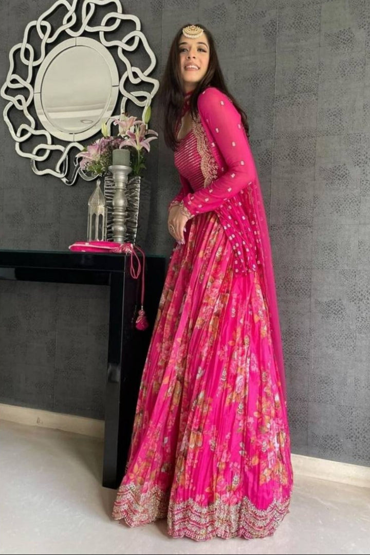 Woman in a bright pink traditional outfit standing in a room with a decorative mirror and table.