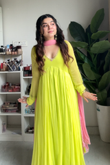 Woman in a bright yellow dress with pink accents standing in a room with shelves and a plant.