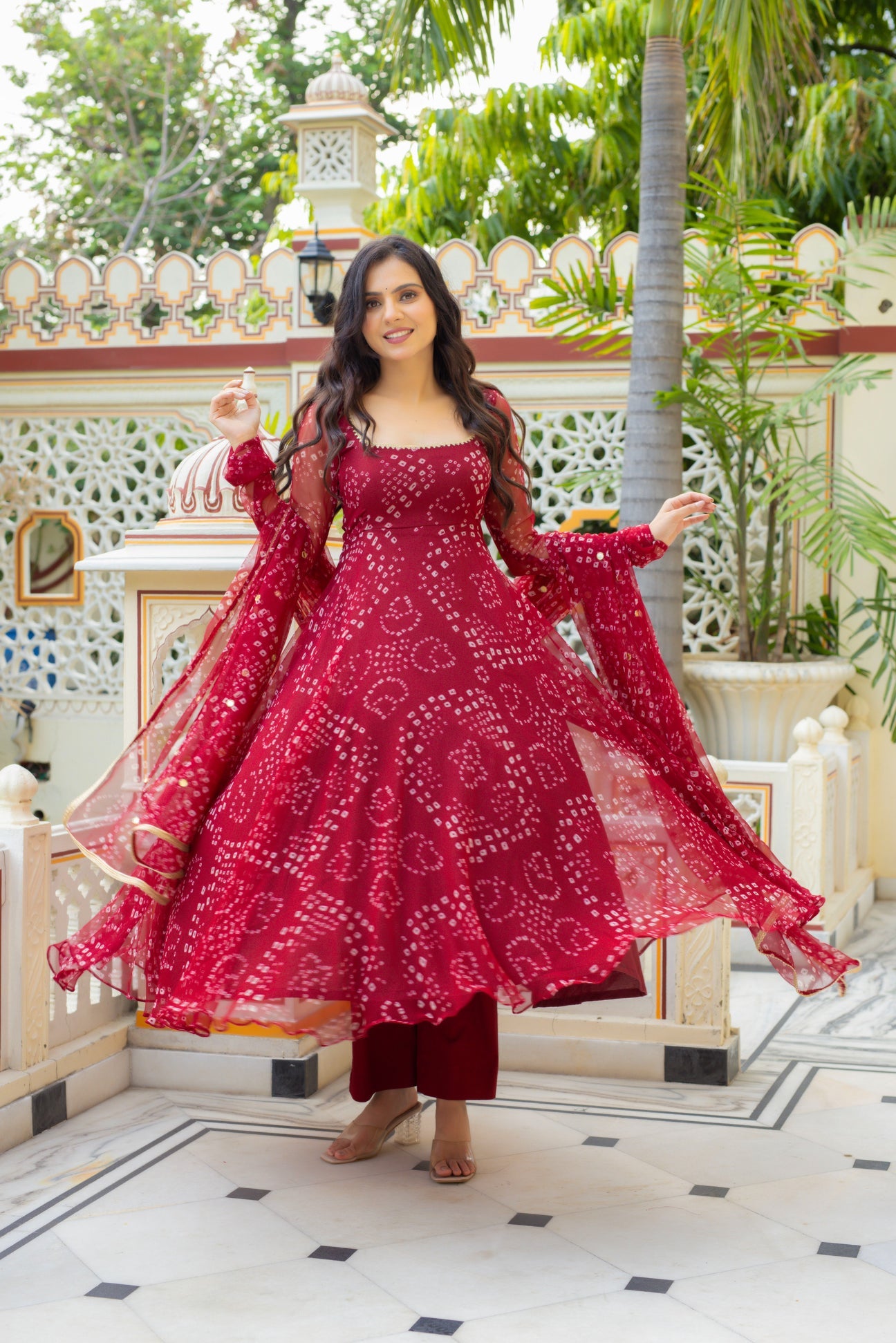 A woman wearing a deep red embroidered Anarkali gown with matching dupatta and maroon pants, standing in a courtyard with white walls and green plants.