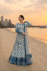 Woman in a blue and white floral dress standing on a beach with a city skyline in the background.