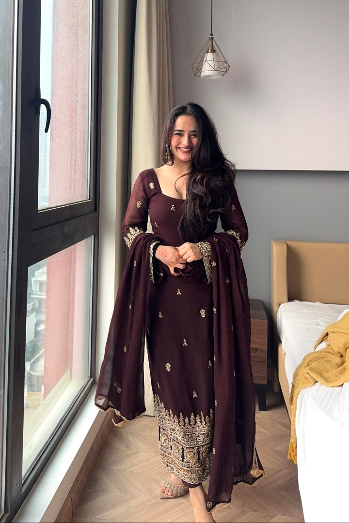 Woman in a dark brown traditional outfit standing by a window in a room with a bed and lamp.