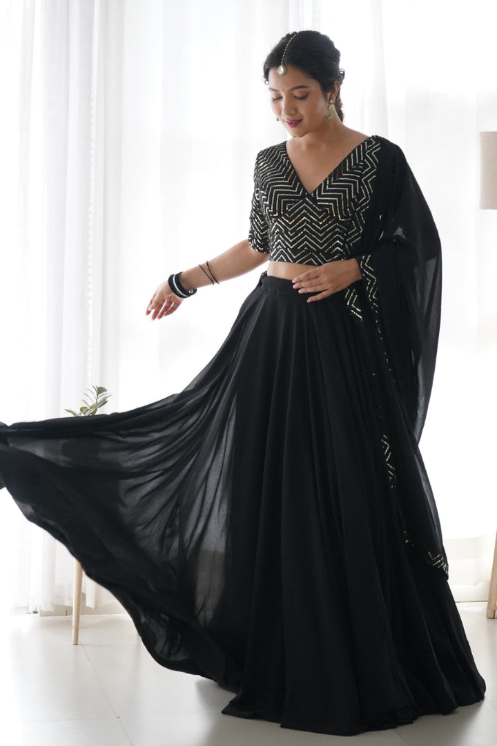Woman wearing a black saree with a white pattern in a room with white curtains and a lamp.