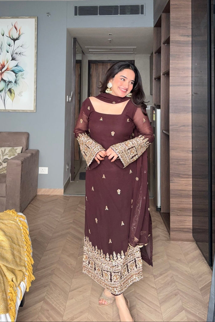 Woman in a brown dress with gold details standing in a room with gray walls and wooden flooring.