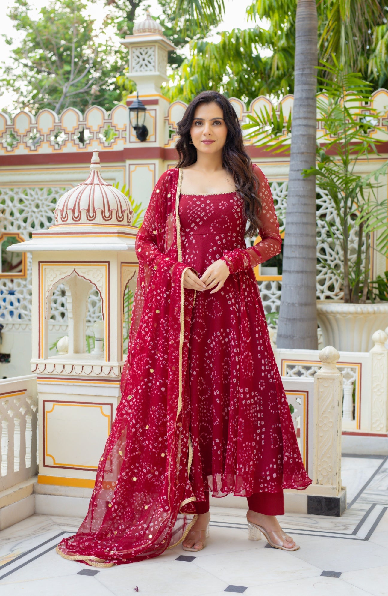A woman models a traditional Indian outfit consisting of a deep red anarkali kurta, matching trousers, and a sheer red dupatta with white prints, set against an ornate courtyard background.