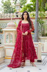 A woman models a traditional Indian outfit consisting of a deep red anarkali kurta, matching trousers, and a sheer red dupatta with white prints, set against an ornate courtyard background.