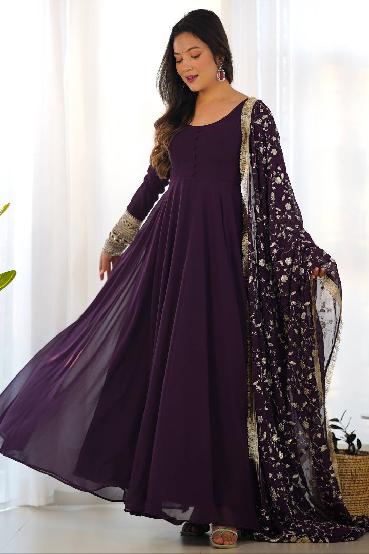 Woman wearing a long purple dress with a floral dupatta against a white curtain background