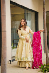 Woman in a beige traditional outfit holding a pink dupatta, standing in front of a glass door.
