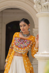 Woman in a traditional yellow and white outfit with intricate designs, standing against a classical architectural background.