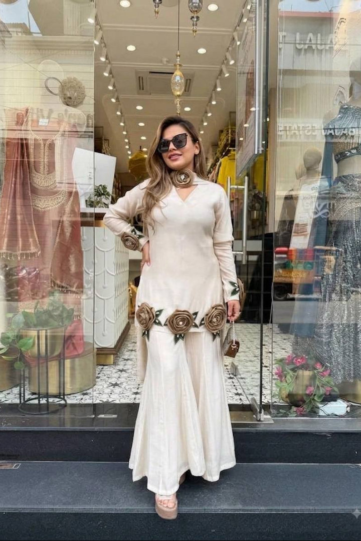 A woman wearing a cream-colored long-sleeve maxi dress with brown floral embellishments stands in front of a clothing boutique window display.