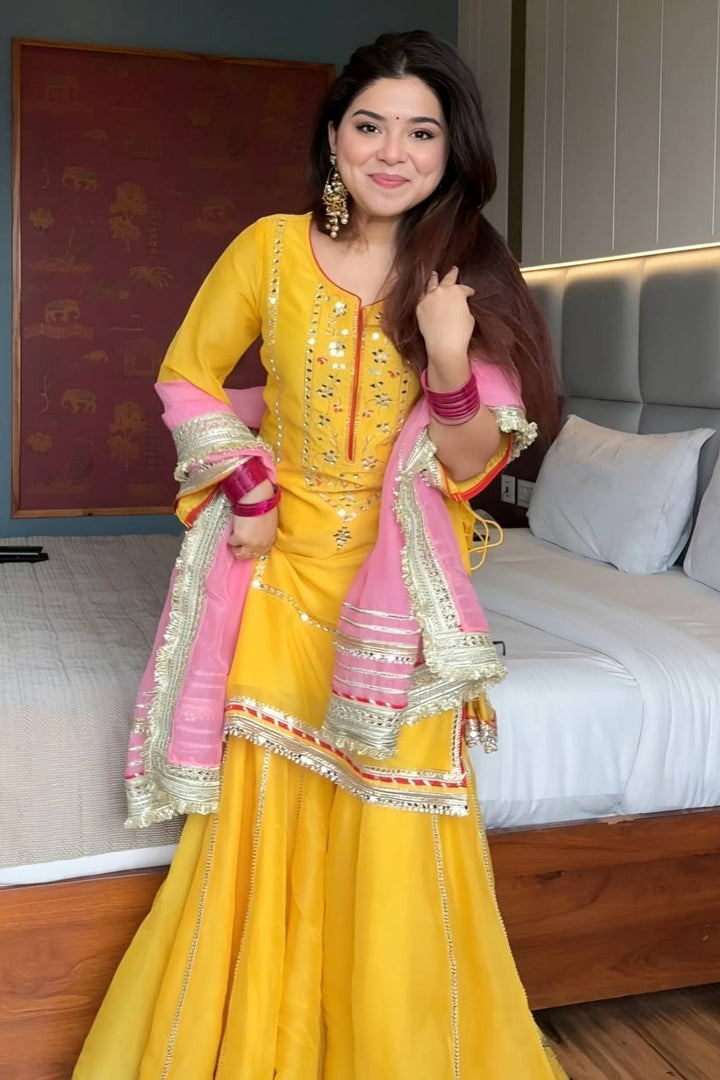 Woman in a yellow traditional outfit with pink and gold accents in a bedroom setting.