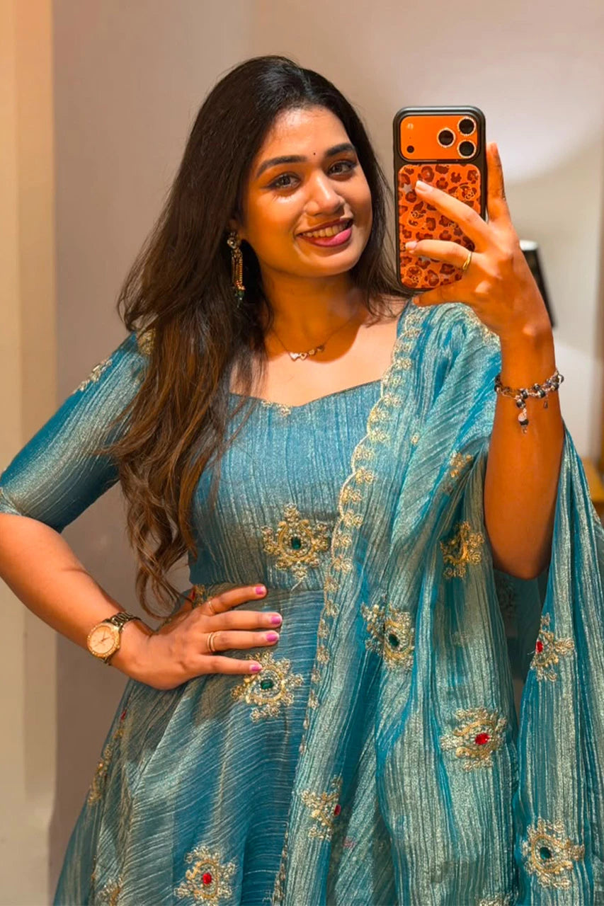 A woman taking a mirror selfie wearing a teal blue embroidered traditional dress with a matching dupatta. The dress features gold floral embroidery and small red and green stone accents.