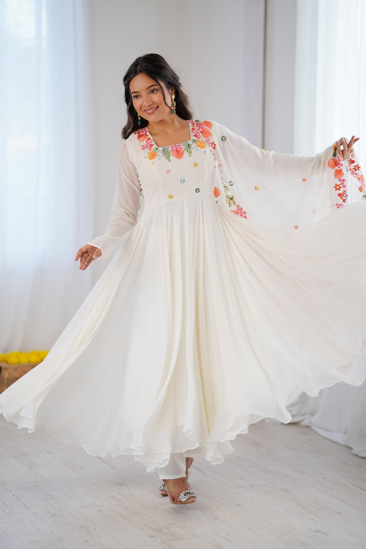 A woman wearing a flowing cream-colored maxi dress with colorful floral embroidery on the bodice and sleeves, twirling in a bright, minimalist room with white curtains.