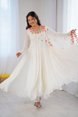 A woman wearing a flowing cream-colored maxi dress with colorful floral embroidery on the bodice and sleeves, twirling in a bright, minimalist room with white curtains.