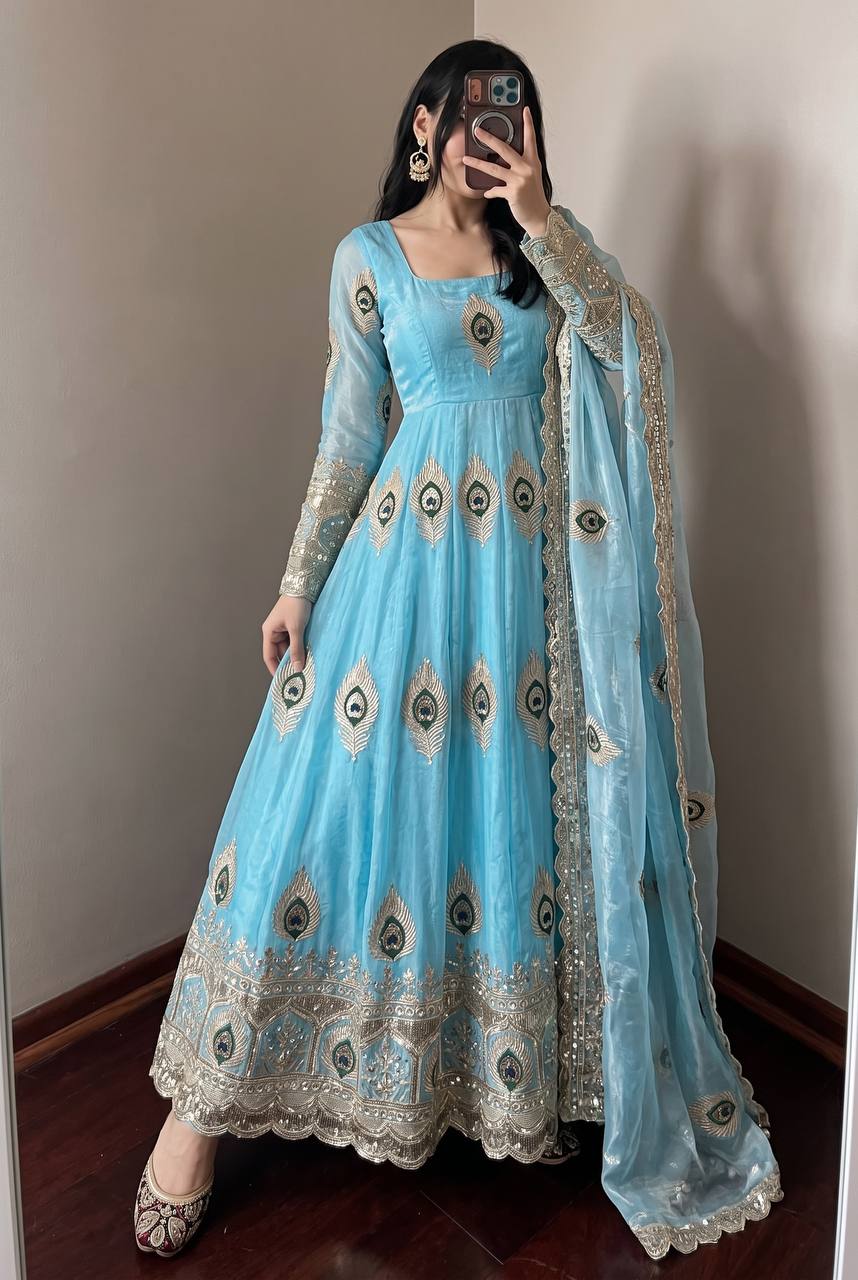 A woman takes a mirror selfie wearing a light blue anarkali gown with intricate gold and silver embroidery, a matching dupatta, and flat shoes in an indoor setting.