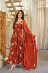 A woman models a rust-orange Anarkali gown with gold floral embroidery and a matching dupatta draped over her arm in an indoor setting with a staircase in the background.