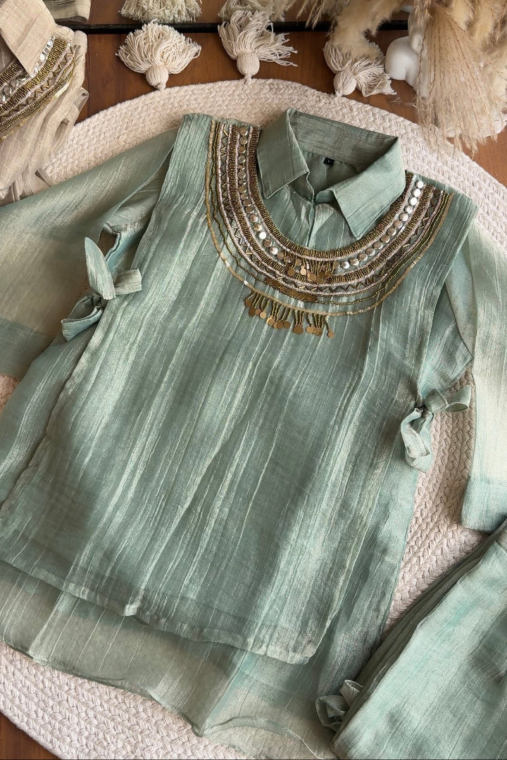 A sage green long-sleeve blouse with intricate gold and silver embroidery around the collar and a tassel detail, laid flat on a woven rug with bohemian decorative props.