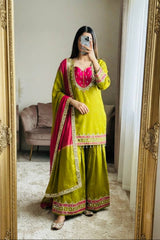 A woman takes a mirror selfie wearing a lime green traditional outfit with a bright pink embroidered neckline and a deep pink dupatta with gold borders. The setting is a home interior with a large gold-framed mirror, a beige sofa, and a white curtain in the background.