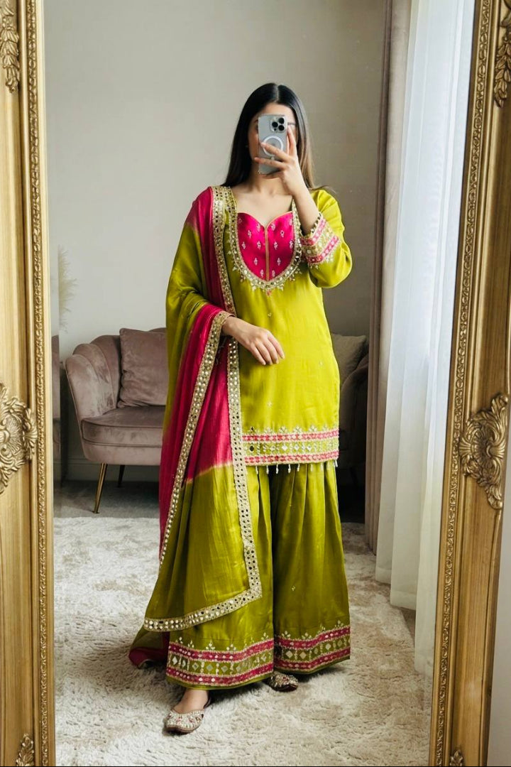 A woman takes a mirror selfie wearing a lime green traditional outfit with a bright pink embroidered neckline and a deep pink dupatta with gold borders. The setting is a home interior with a large gold-framed mirror, a beige sofa, and a white curtain in the background.