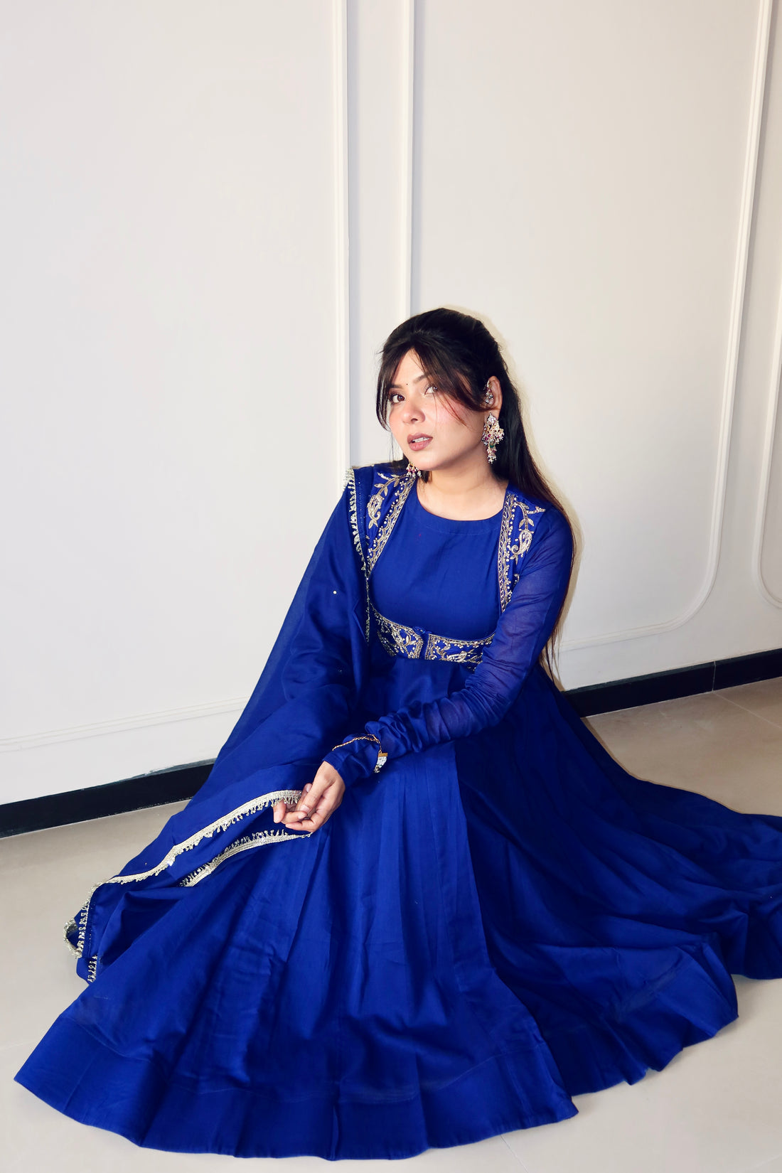 A woman wearing a royal blue traditional gown with silver embroidery sits on a light-colored floor against a white paneled wall.