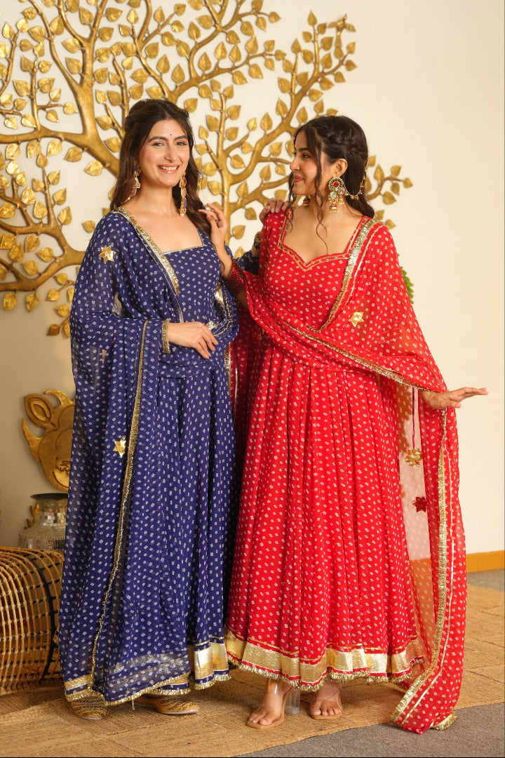 Two women modeling traditional Indian Anarkali gharara suits in a decorated indoor setting. One suit is deep blue with white polka dots and gold borders, and the other is red with white polka dots and gold borders.