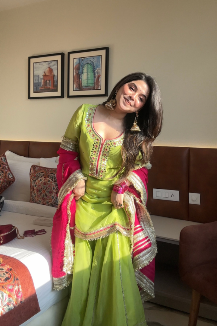 Woman in a green and pink traditional outfit standing in a room with a bed and wall art.