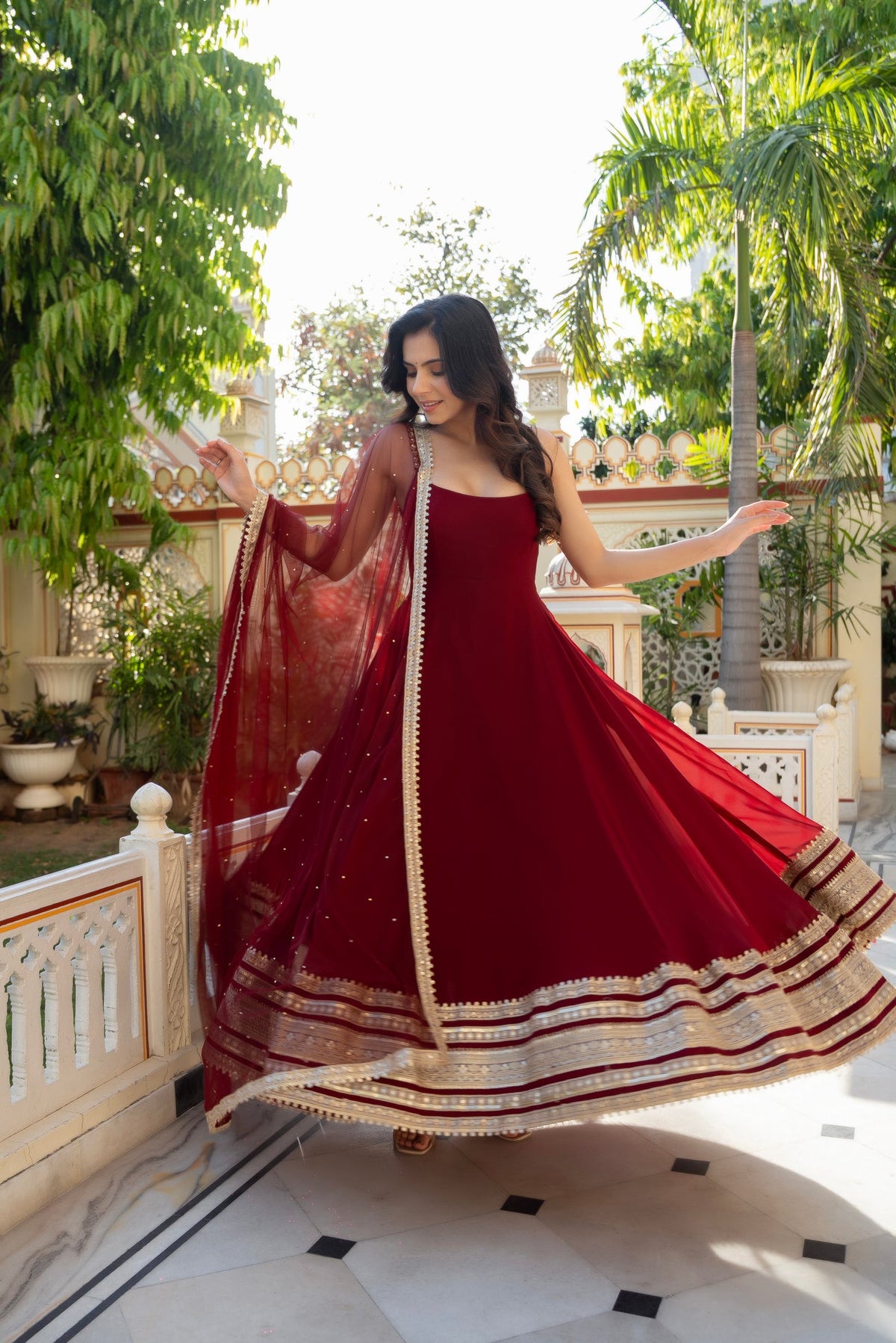 A woman wearing a deep red anarkali gown with gold border detailing twirls outdoors on a tiled patio surrounded by green plants and white railings.
